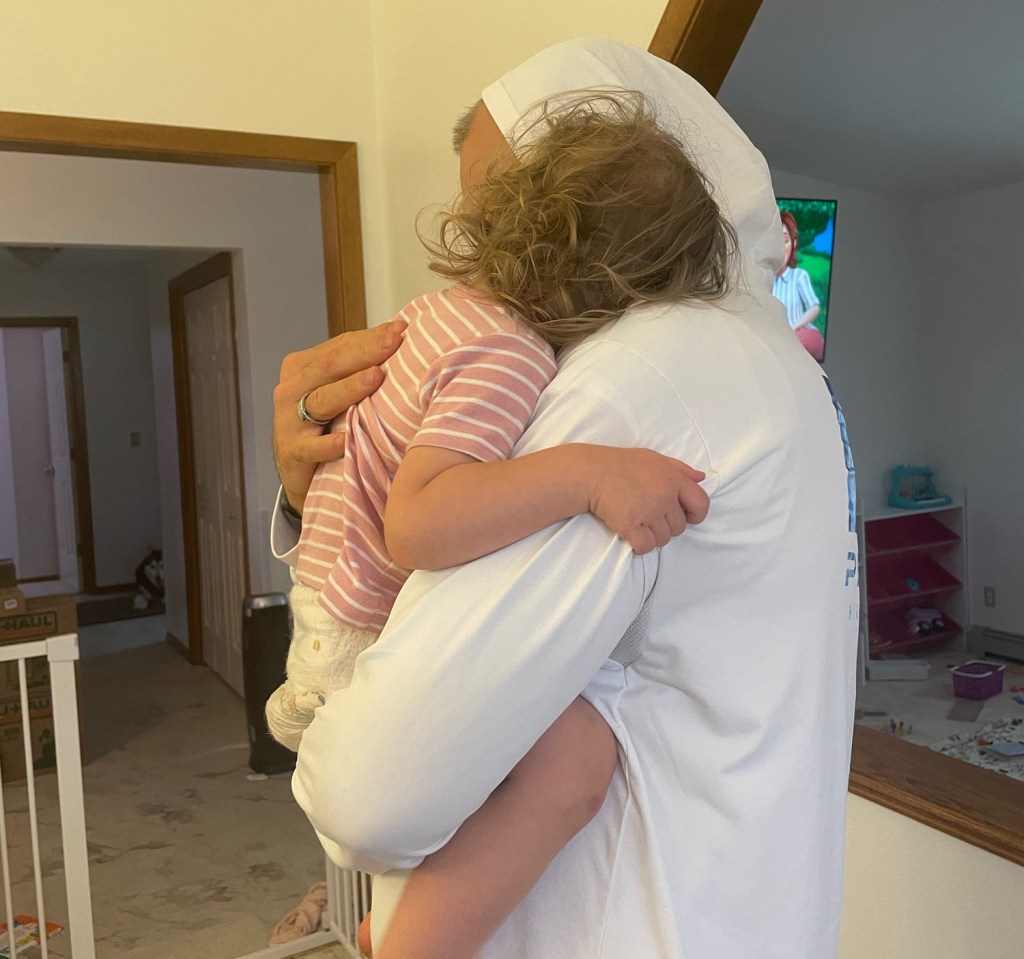 A man in a hooded shirt holds a toddler, who is clasping him tightly around the neck. They are inside a home near an open doorway leading to another room where a TV shows a nature scene.