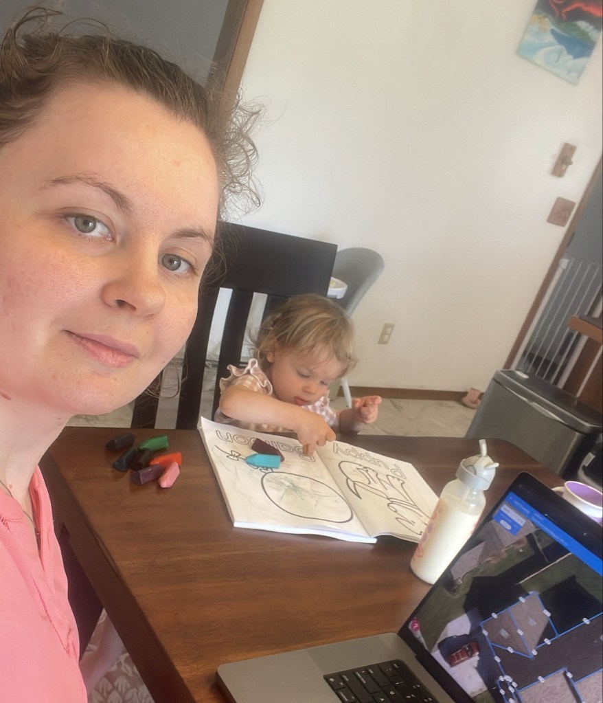 A woman with a slight smile looks at the camera, sitting beside a toddler who is focused on coloring at a dining table. In front of the child are various crayons and a coloring book, with a laptop by them. 