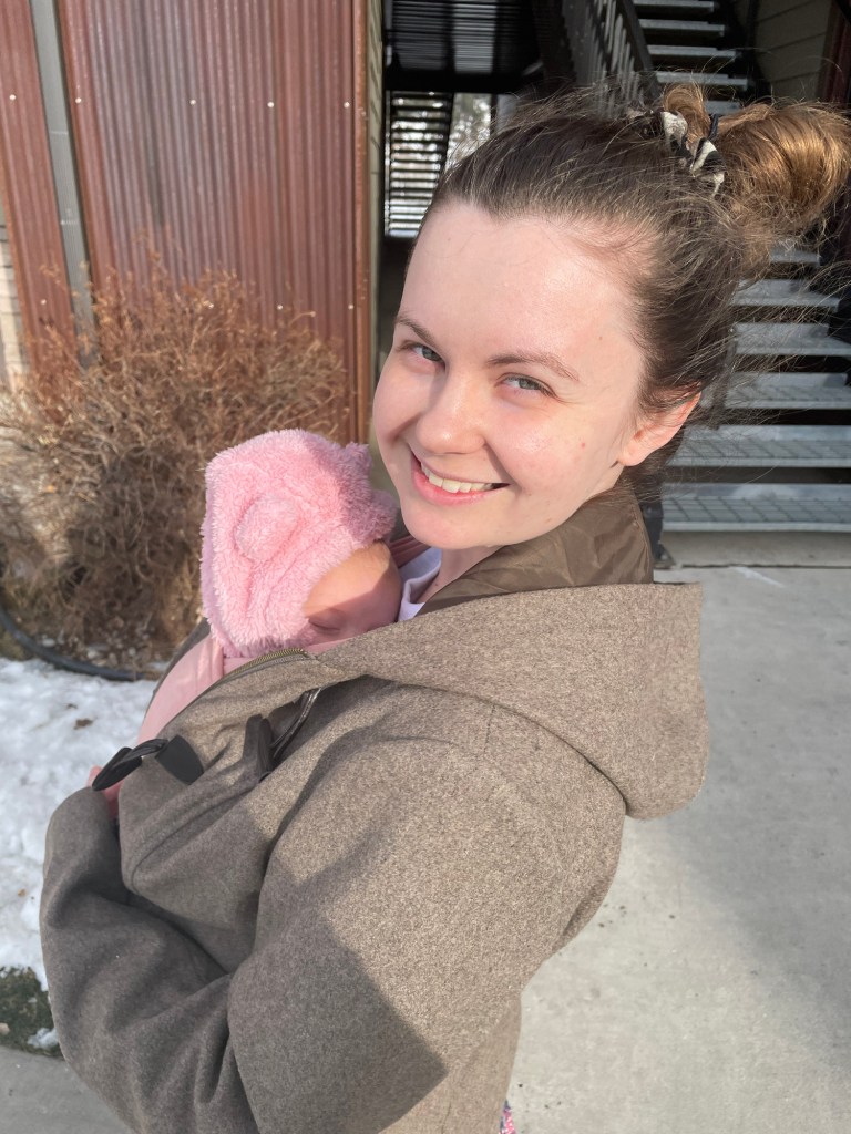 A smiling woman holds a young child wrapped in a pink blanket, standing outside on a sunny day, with winter vegetation and a metal structure in the background.