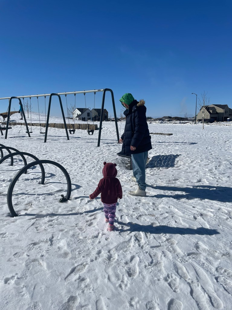 A toddler in a pink snowsuit and an adult in a blue coat and green beanie walk on a snowy playground with swings and play structures, under a clear blue sky.