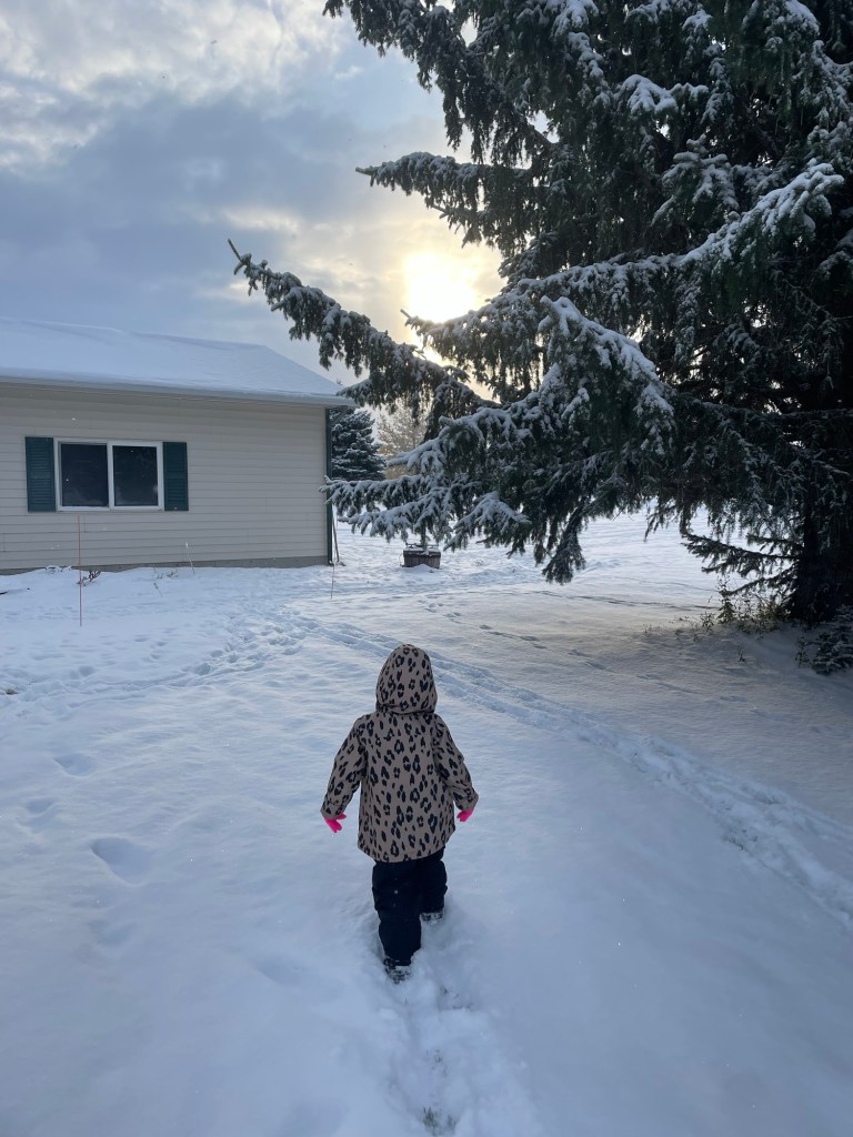 A small child dressed in a leopard print jacket and dark pants runs joyfully through a snowy landscape, with snow-dusted pine trees and a house in the background, highlighted by the soft winter sunlight breaking through the clouds.