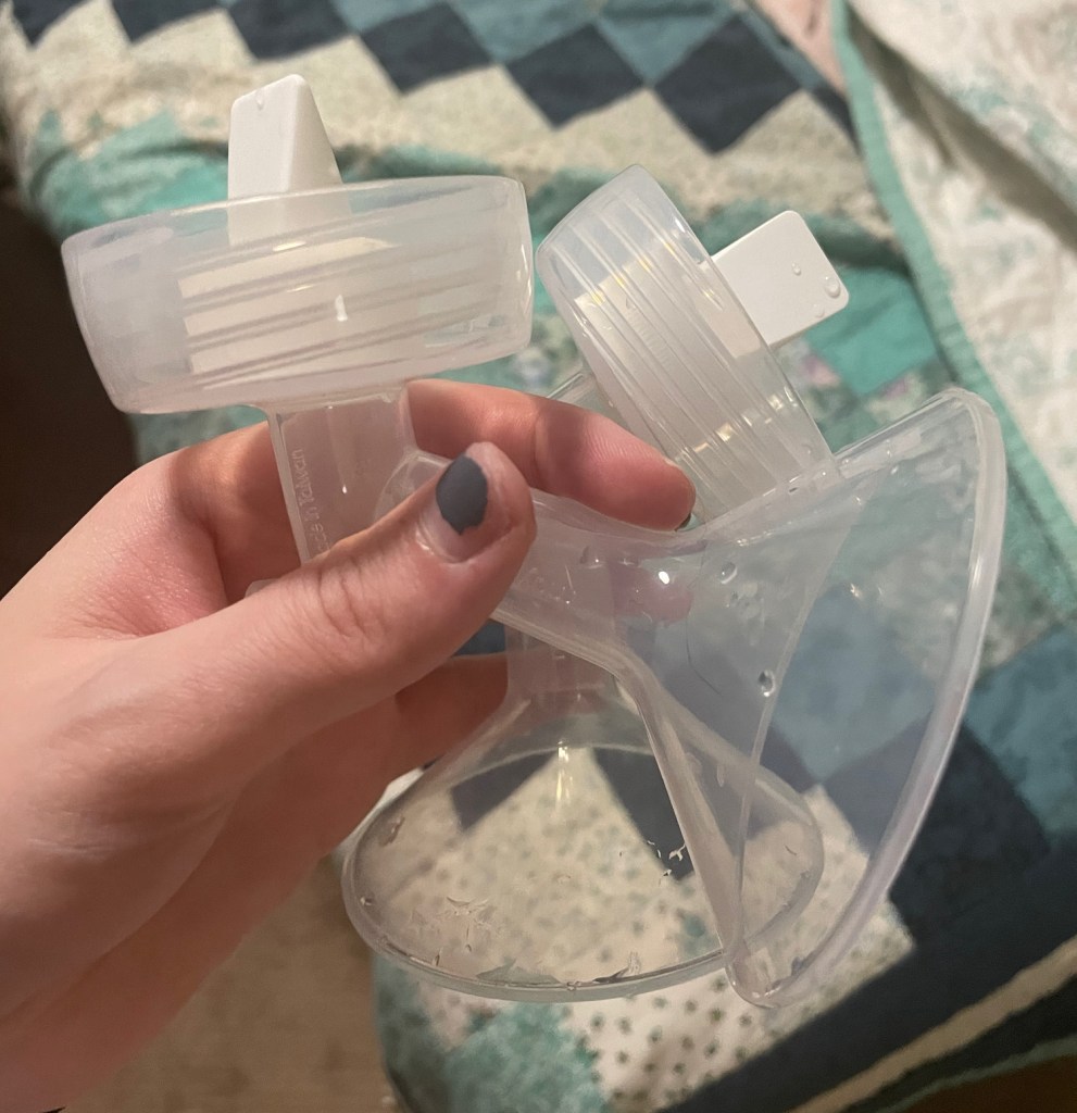 a close-up of a hand holding one of the breast pump's flanges, showing its structure including the attached valve and membrane, which are essential parts for its suction and release mechanism. This image shows a  quilt with a blue and green floral pattern in the background 